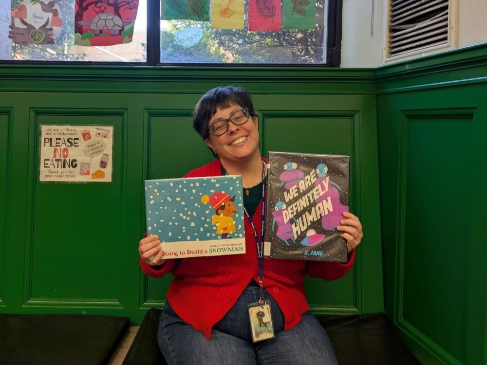 Woman with glasses and short hair smiling holding up 2 children's books