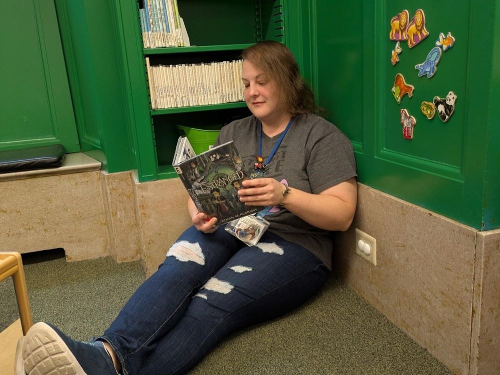 woman sitting on ground with bookshelf behind her reading children's book