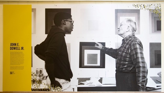 a young John Dowell talking to German-American artist Josef Albers in front of a wall covered with Albers prints