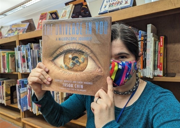 Person wearing mask holding up a children's picture book in front of bookshelves
