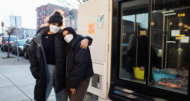 Two masked people standing with arms around each other next to The People's Fridge outside a library