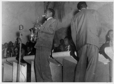 The Jimmy Heath Orchestra performing in 1947, Jimmy conducting, Charlie Parker sitting in, and John Coltrane between them, watching his hero wide-eyed
